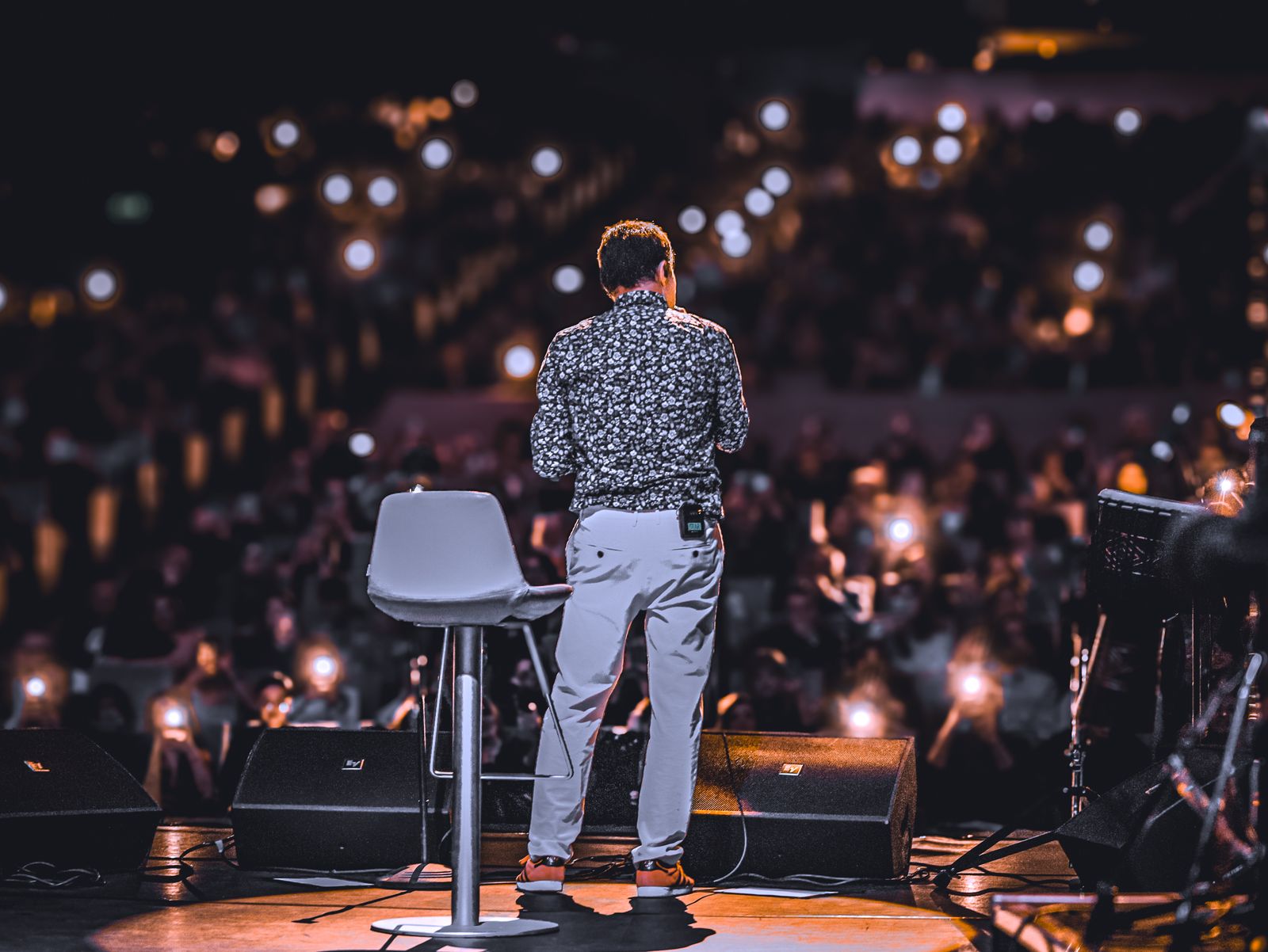 Palestrante visto de costas no palco diante de auditório lotado com luzes de celular acesas, cena típica dos palestrantes mais cotados do Brasil