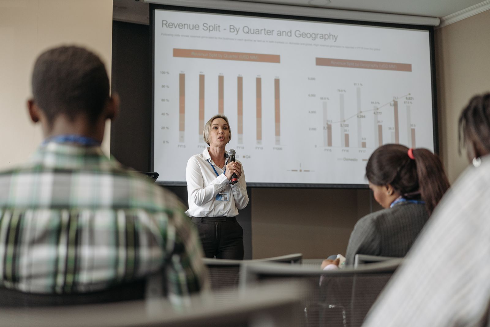 Palestrante com microfone apresentando gráficos de receita em tela de projeção para plateia de executivos sentados, formato típico de palestra sobre reforma tributária nas empresas